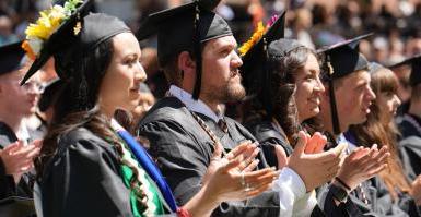 一排戴着黑色帽子、身穿黑色长袍的毕业生站着鼓掌, 在他们身后可以看到一排排的学生和家庭成员.